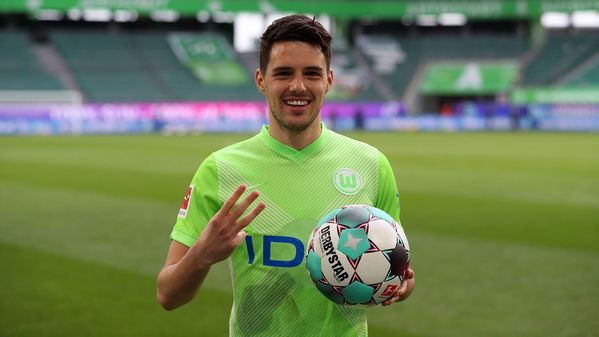 VfL-Wolfsburg-Spieler Josip Brekalo hält in der einen Hand den Ball, mit der anderen zeigt er drei Finger hoch und läschelt in die Kamera.
